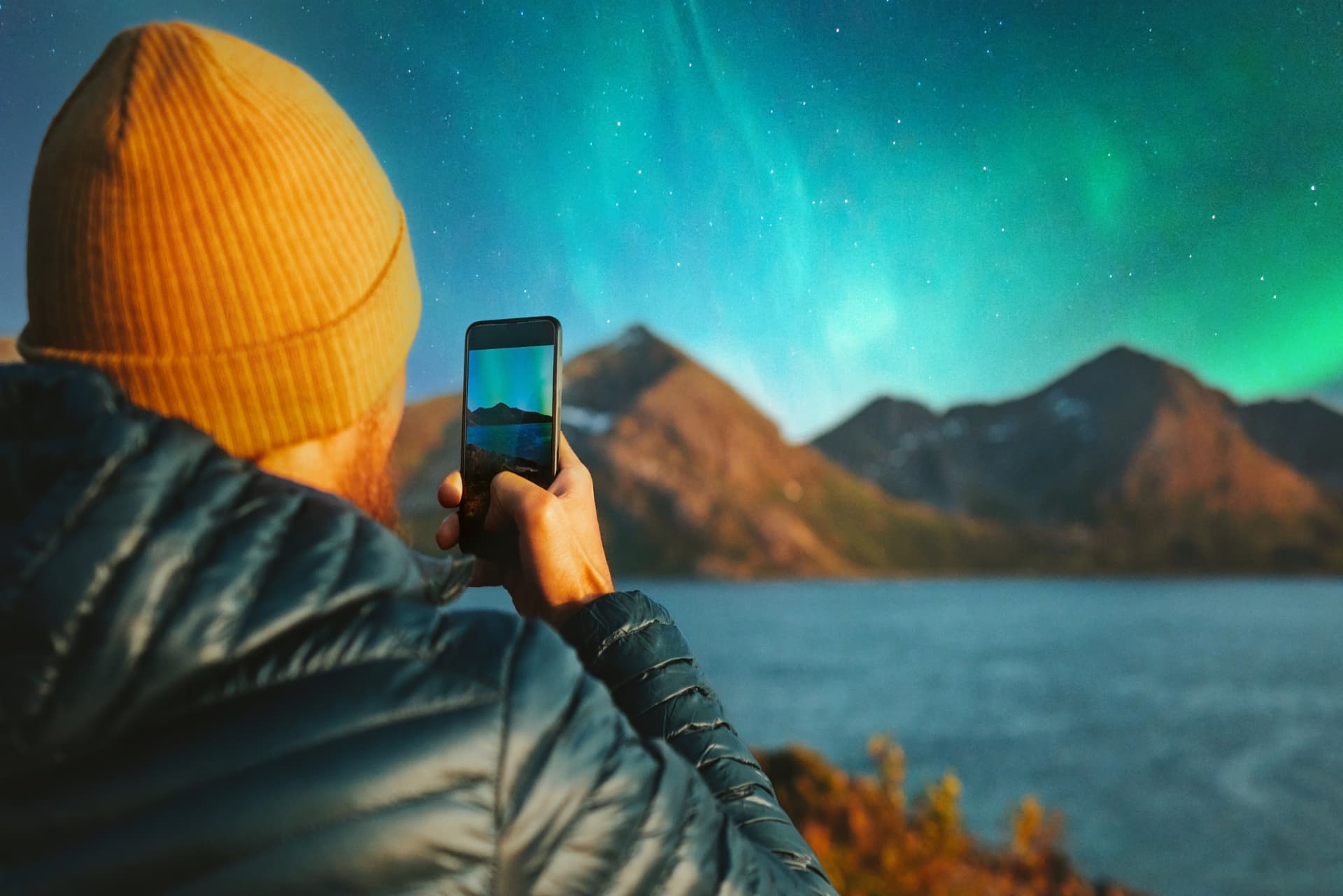 Traveller in an orange beanie photographing the Northern Lights over mountains and a fjord with a smartphone