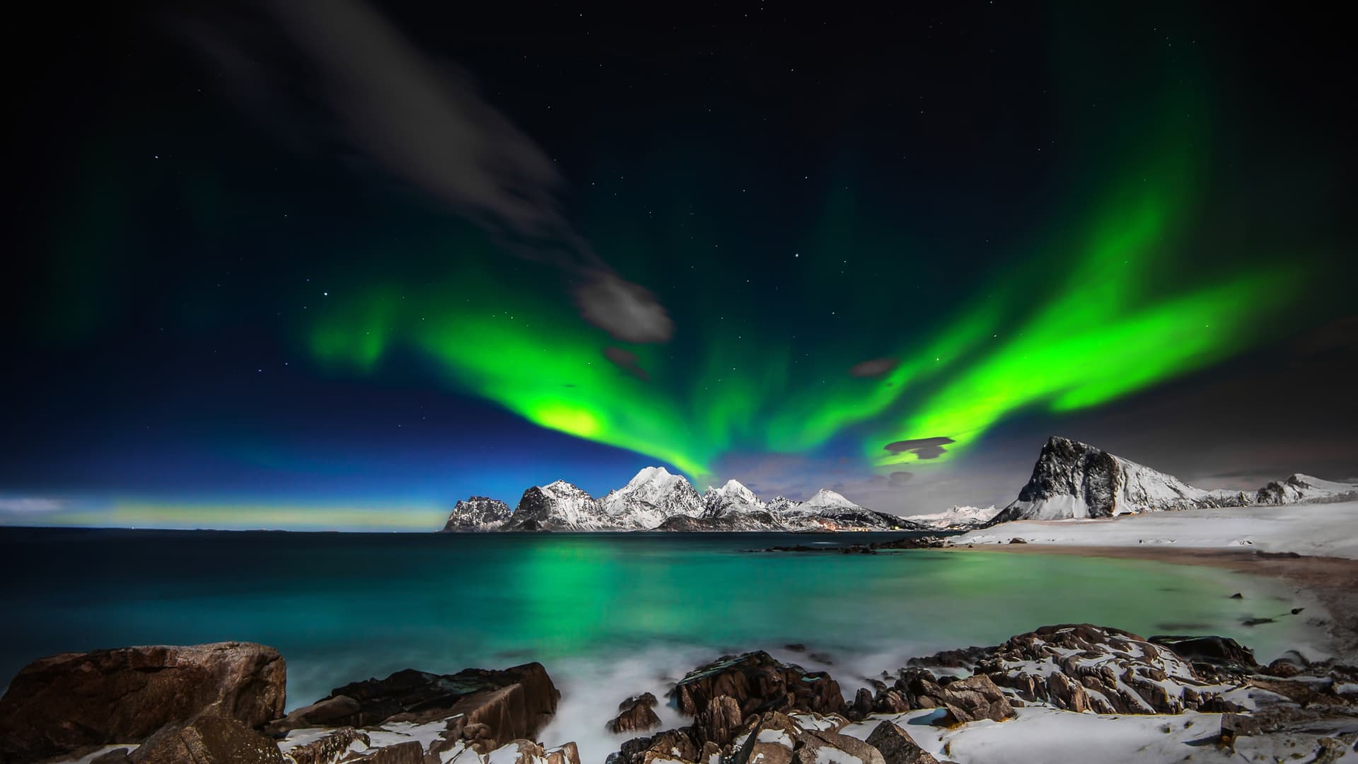 Green aurora over a snow-covered Arctic coastline with mountains and still water